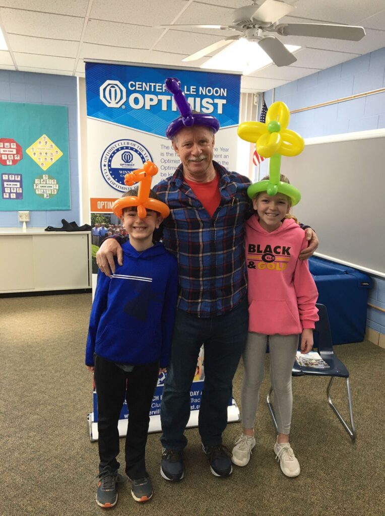 3 people, a boy, a girl, and an Optimist member standing in front of a blue Centerville Noon Optimist sign and wearing balloon hats