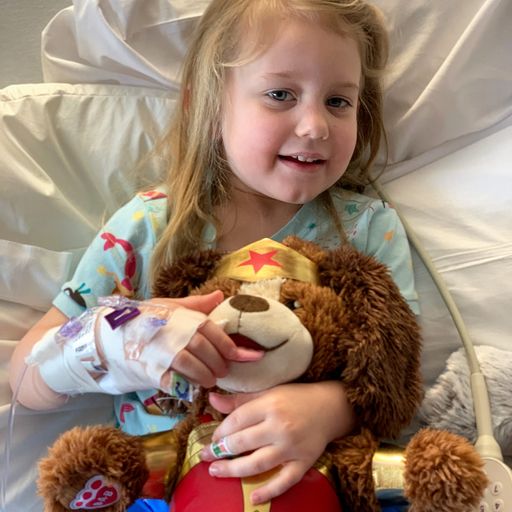 A young girl in a hospital bed holding a Build-A-Bear dog in a Wonder Woman costume